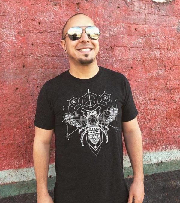 A man smiling wearing sunglasses and a black t-shirt with a geometric bee design.